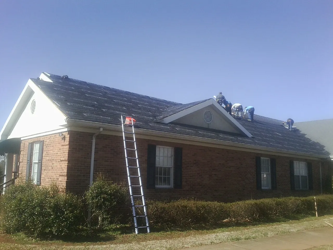Crew tearing shingles off a brick ranch roof with a ladder set — Emergency Roof Repair work by our crew in Marquette